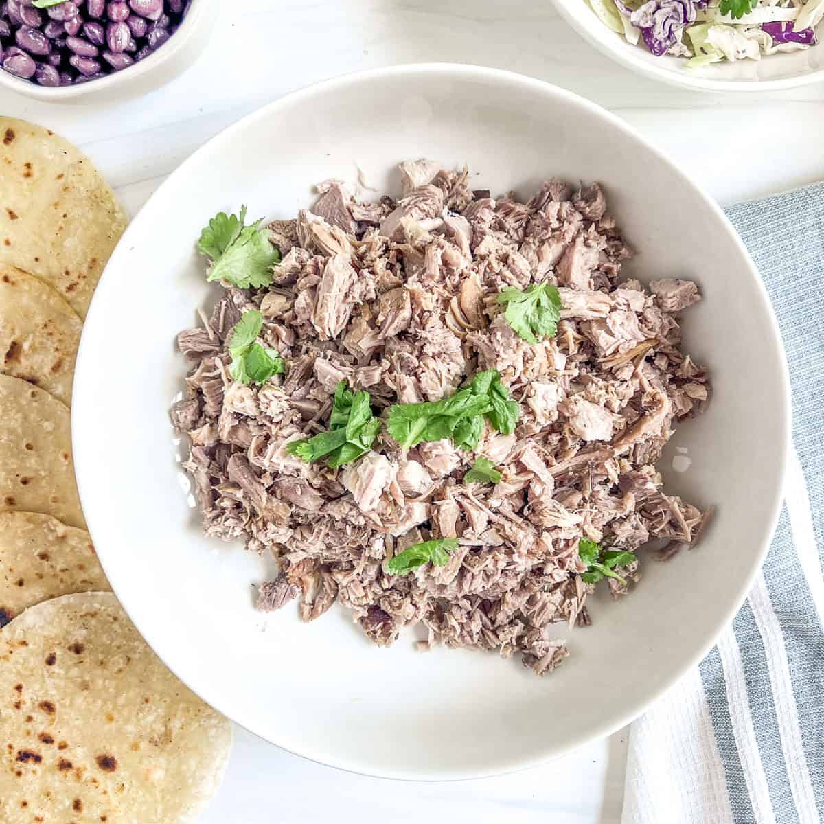 cooked pork meat chopped up in small pieces with fresh cilantro on top with corn tortillas to the side and a bowl of black beans and a bowl of cole slaw to the side