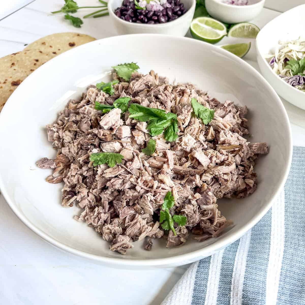 cooked pork meat chopped up in small pieces with fresh cilantro on top with corn tortillas to the side and a bowl of black beans and a bowl of cole slaw to the side and cut up limes to the side
