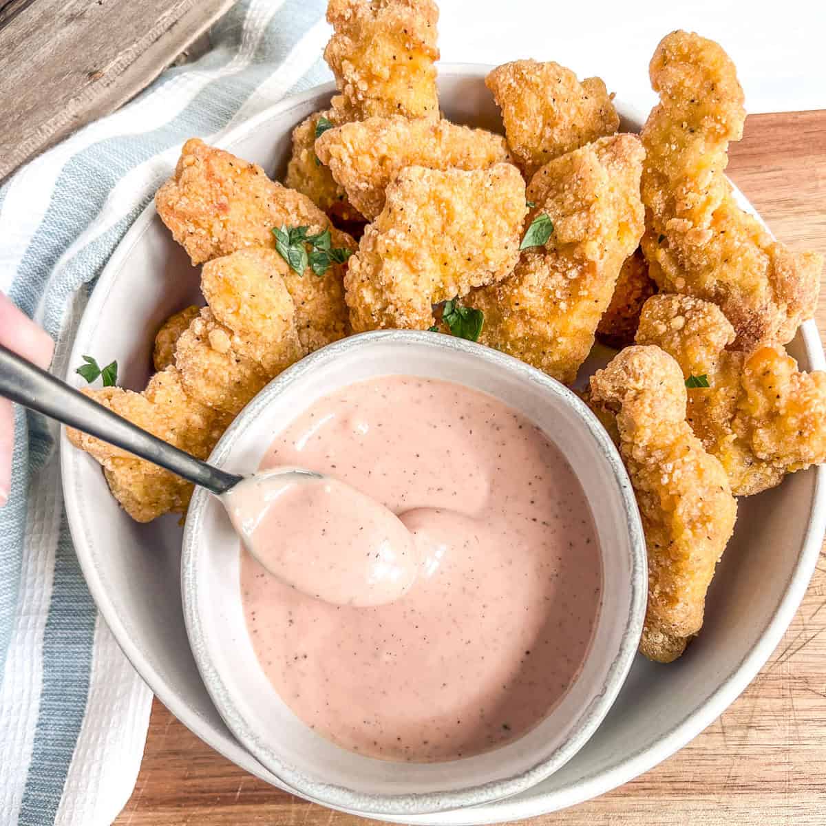 hand holding a spoon of chicken sauce and chicken tenders around the bowl of sauce
