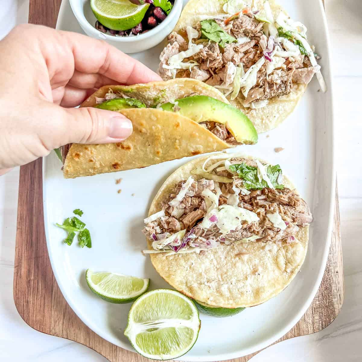 hand holding a taco with slices of avocado and pork meat inside the taco and tacos on the plate