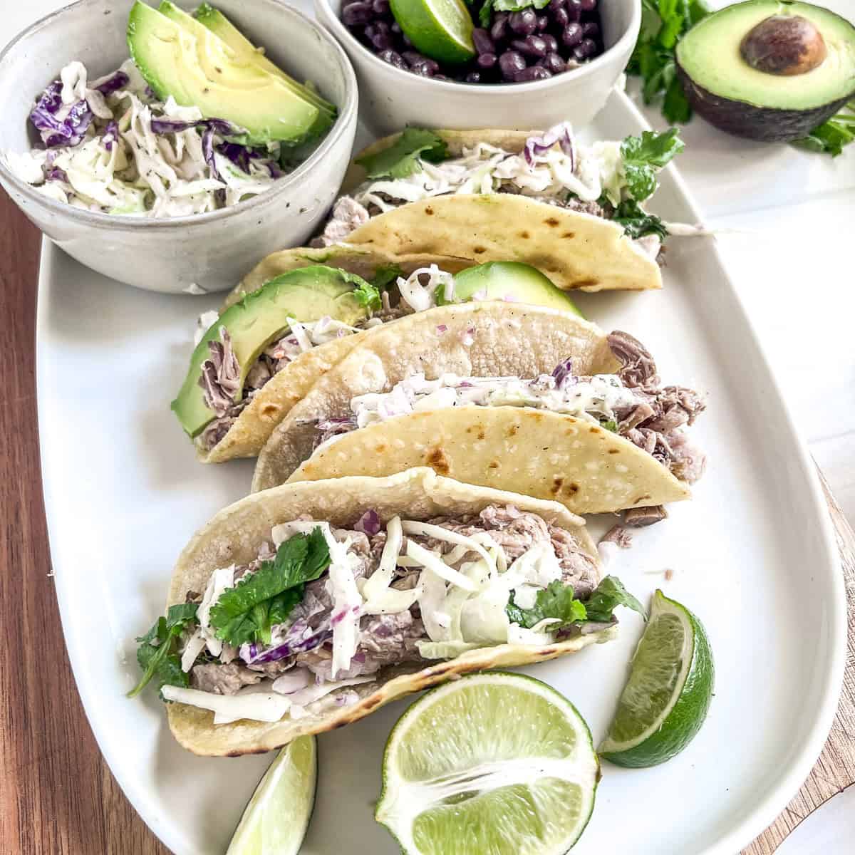 side angle picture of tacos on a white plate with pork, cabbage, cilantro, and avocado slices on top and a bowl of cole slaw to the side