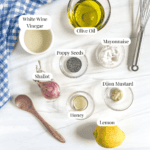 bowl of olive oil, white wine vinegar, whole shallot, bowl of poppy seeds, bowl of honey, bowl of dijon mustard, bowl of mayonnaise, whole lemon, a whisk and a wooden spoon to the side